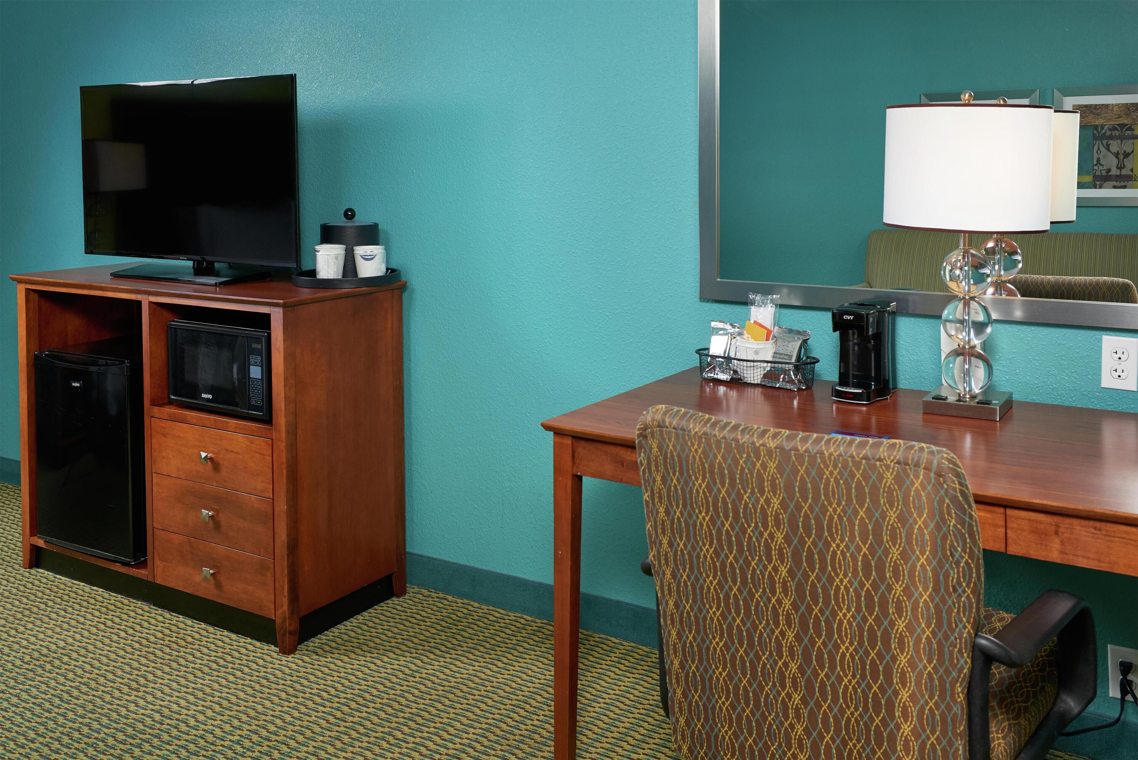 A hotel room with a wooden desk, a television, a minibar, and a patterned armchair.