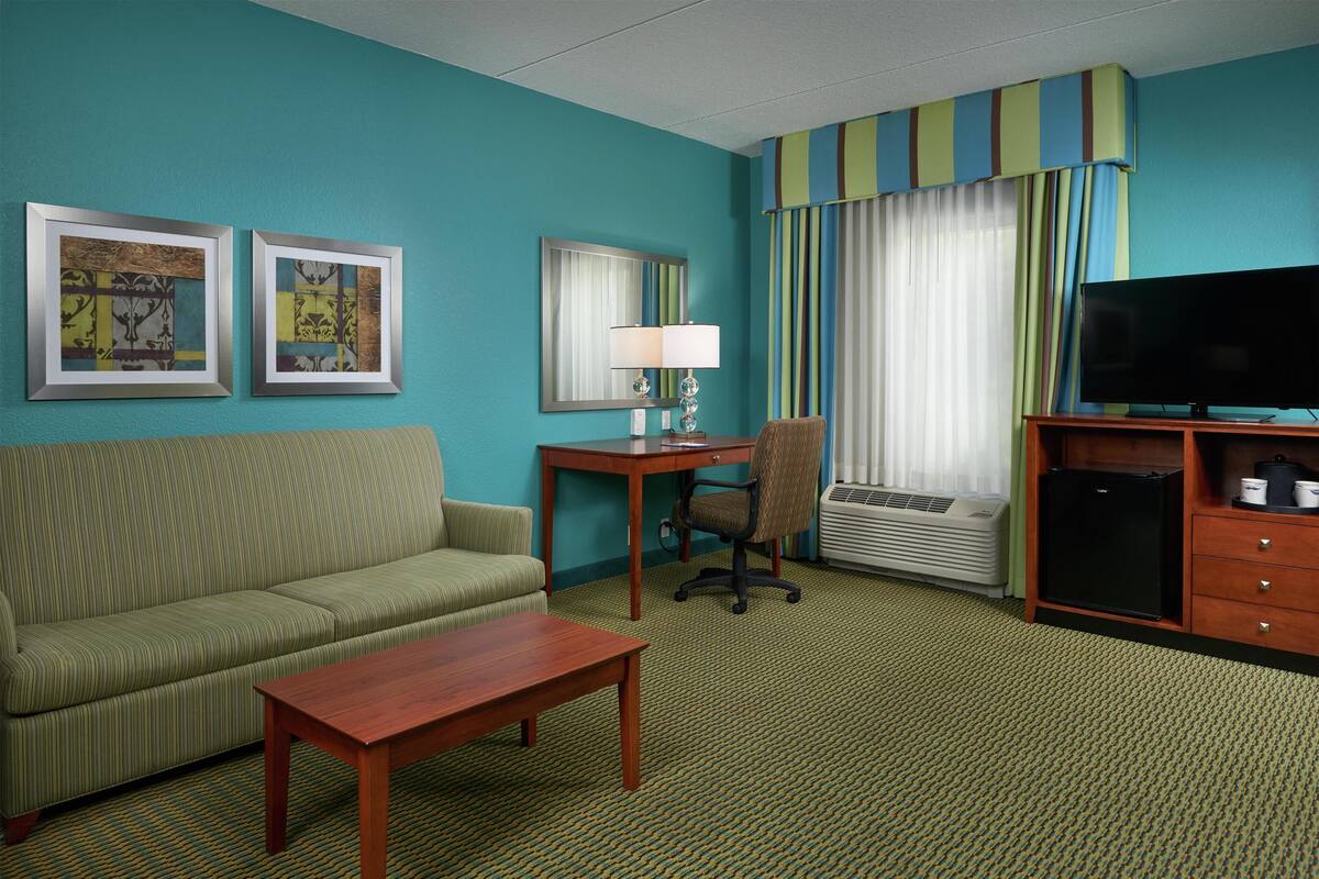 A hotel room with a green sofa, a wooden coffee table, a desk with a lamp, a TV, and a window with curtains.