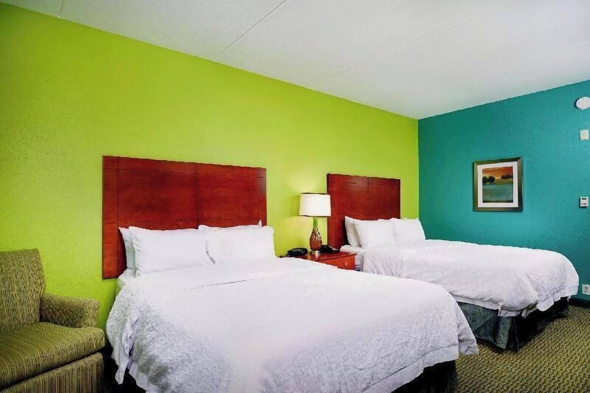 A hotel room with two beds, a green wall, a red headboard, a green armchair, and a painting on the wall.
