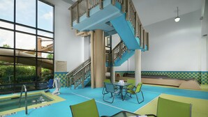 An indoor swimming pool area with a glass-enclosed pool, a blue staircase, and green chairs around a table.
