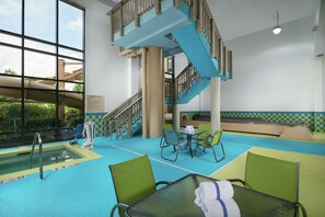 An indoor swimming pool area with a glass-enclosed pool, a blue staircase, and green chairs around a table.