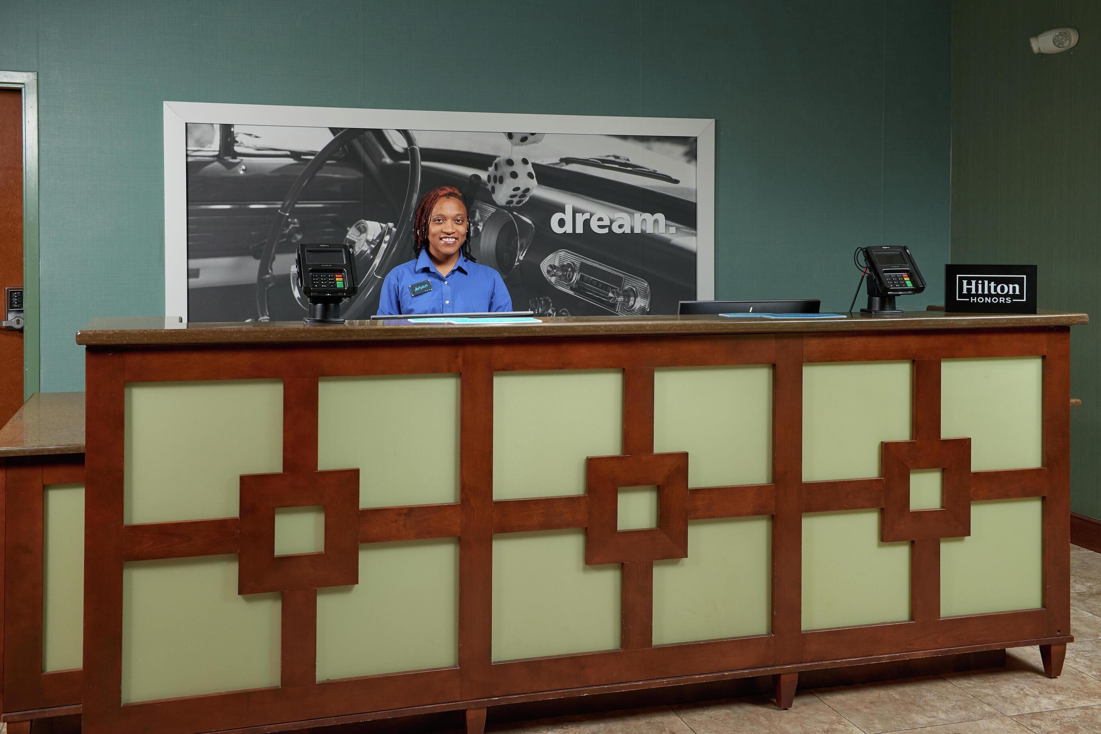 A reception desk with a large poster in the background, a digital device on the counter, and a sign indicating the hotel name.