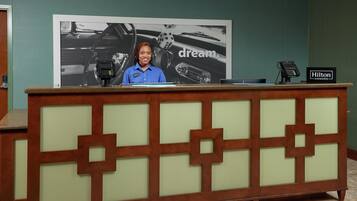 A reception desk with a large poster in the background, a digital device on the counter, and a sign indicating the hotel name.
