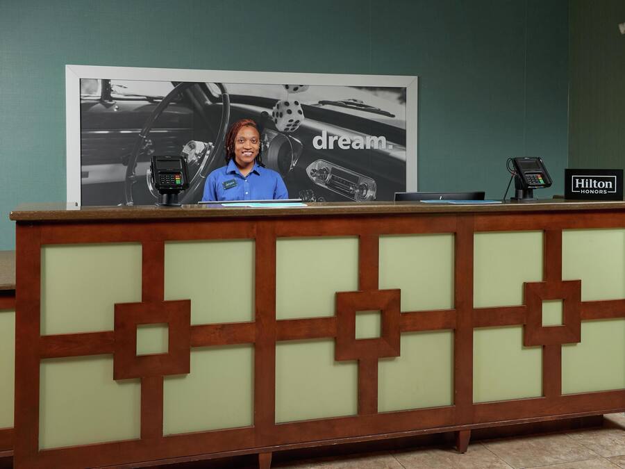 A reception desk with a large poster in the background, a digital device on the counter, and a sign indicating the hotel name.