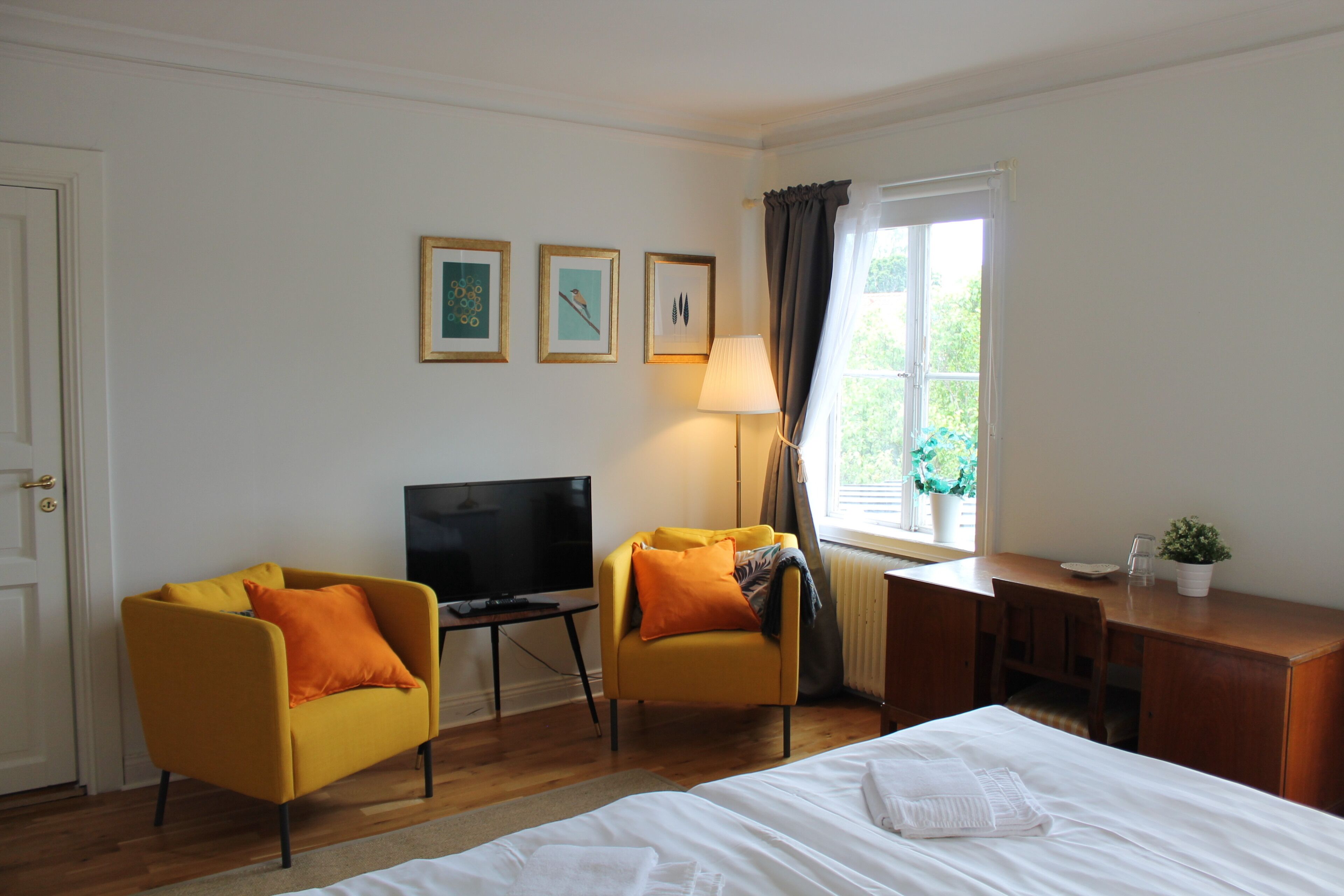Habitación de hotel con cama, dos sillones amarillos, escritorio con computadora, televisor y ventana con cortinas.