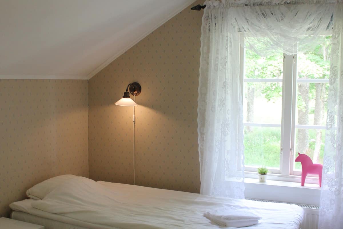 A small, single-bed room with a sloped ceiling, a window with a view of greenery, a wall-mounted light, and a small table with a chair.