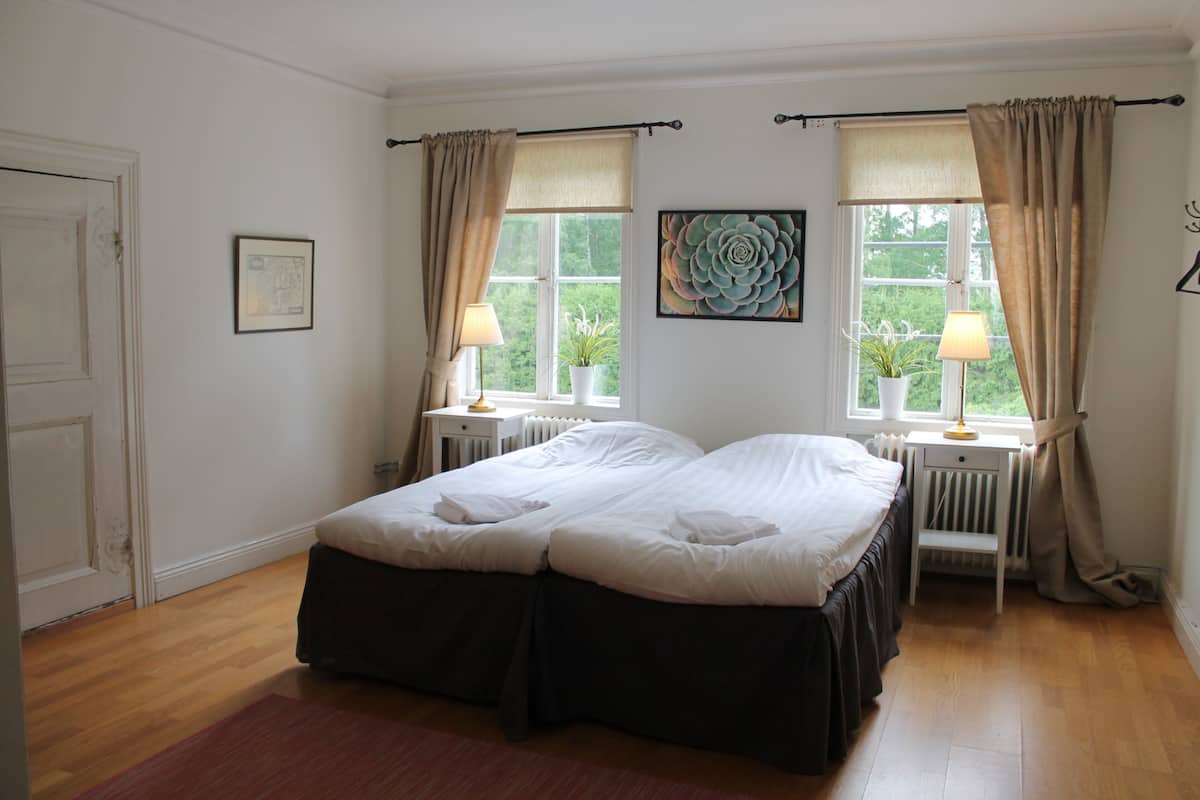 A double bed with a dark skirted base, two bedside tables with lamps, a radiator, and a window with curtains.