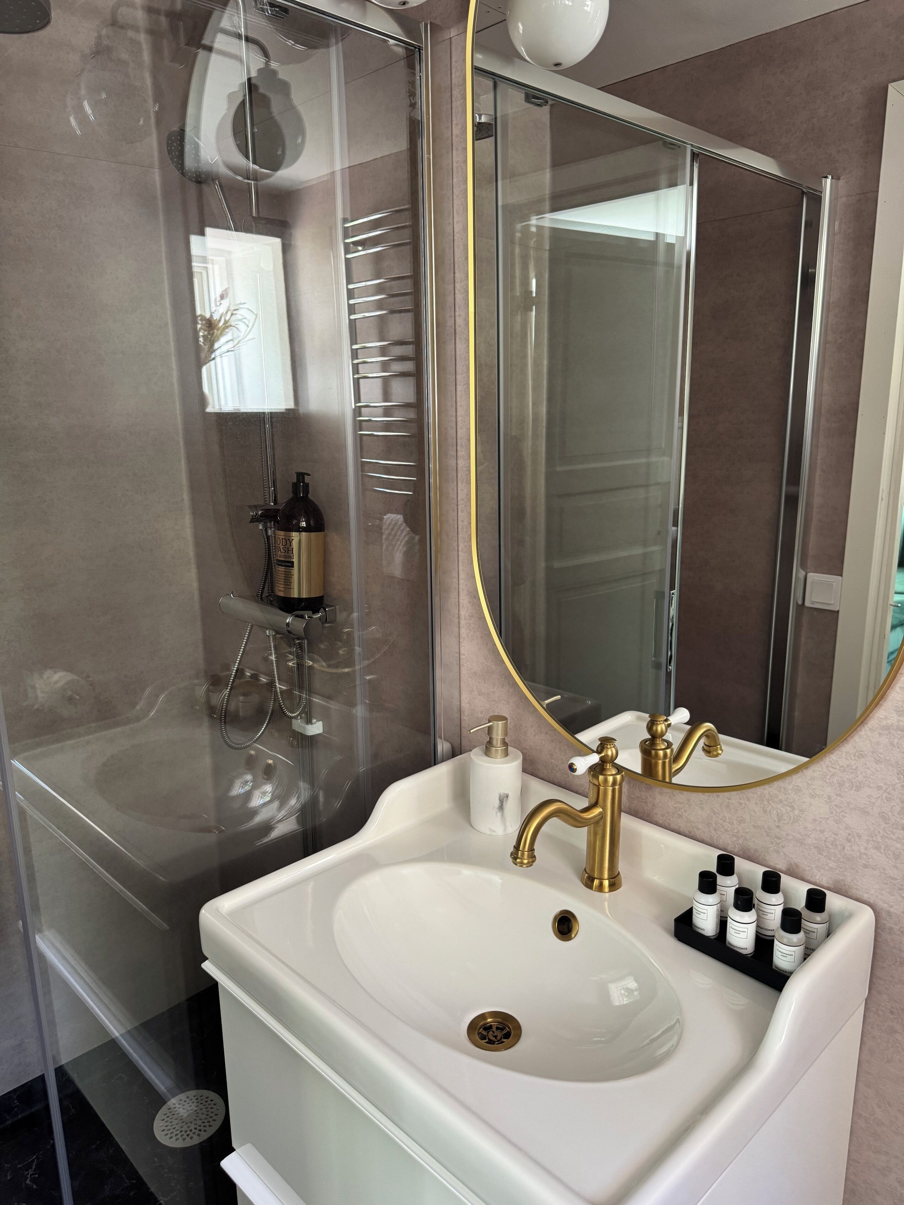 A bathroom with a glass-enclosed shower, a white sink with gold fixtures, and a large mirror.