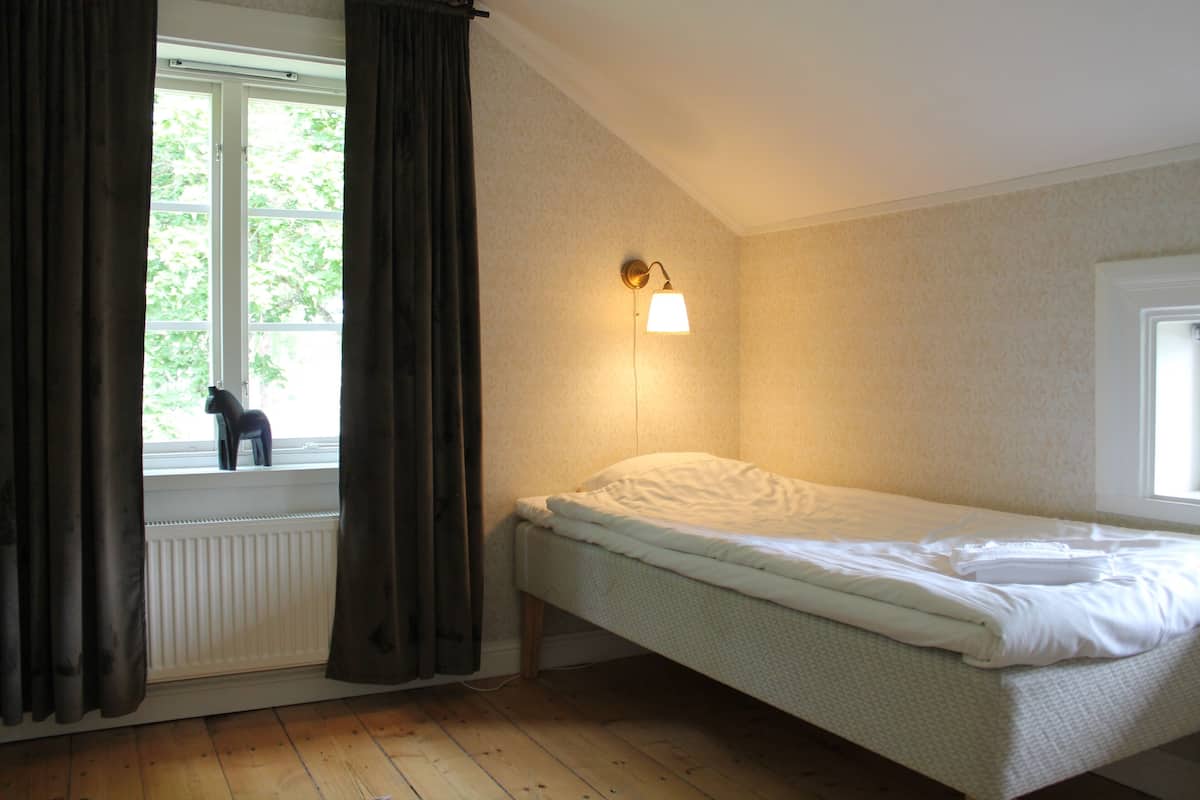 A single bed in a room with a window, a radiator, and a wall-mounted light fixture.