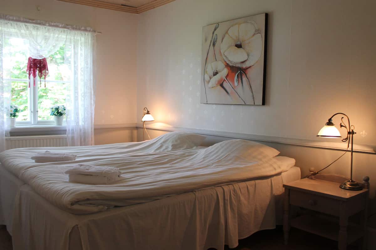 A neatly made bed with folded towels, a bedside table with a lamp, and a window with sheer curtains.