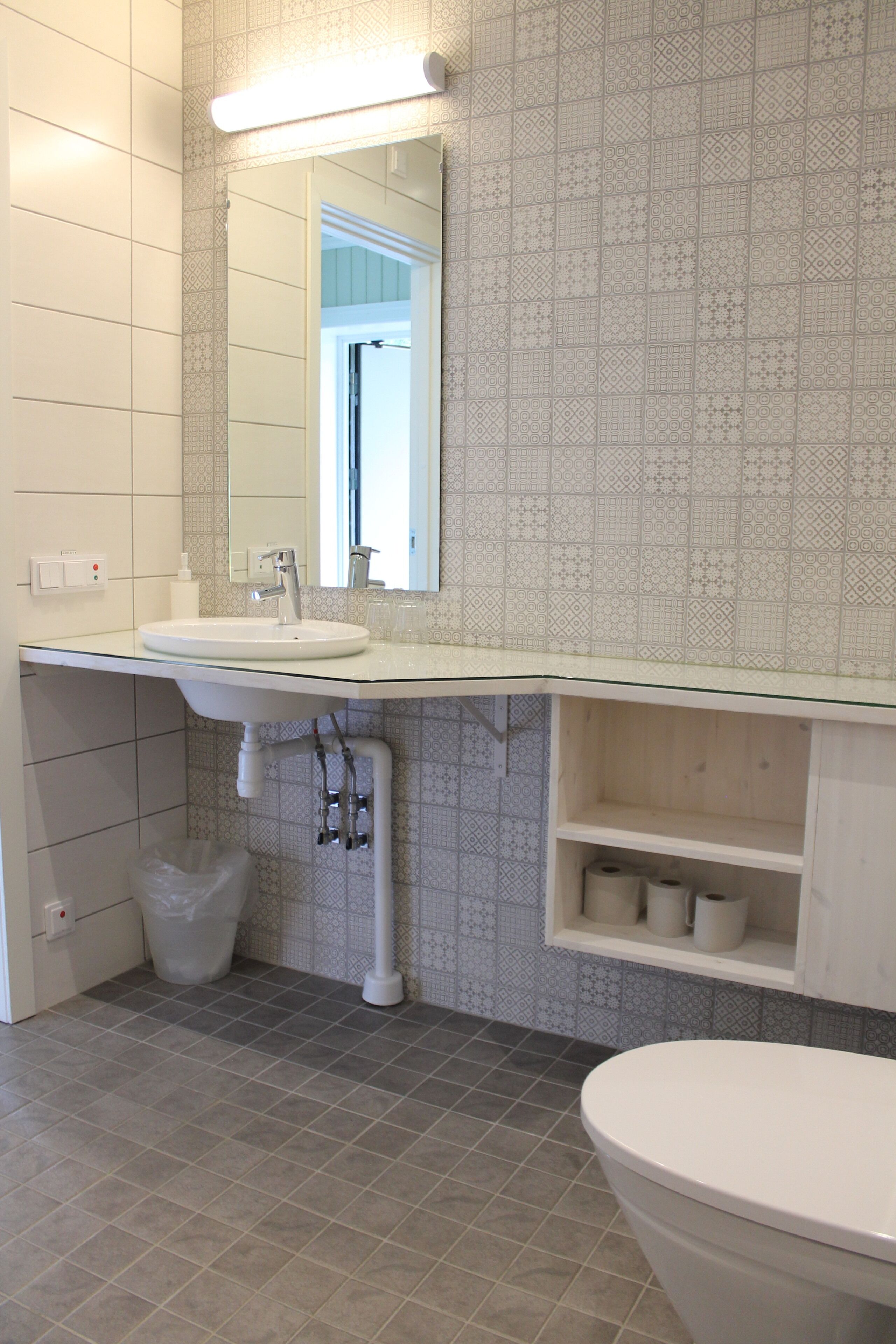 A bathroom with a white sink, mirror, and toilet. There is a patterned tile backsplash and a shelf with toiletries.