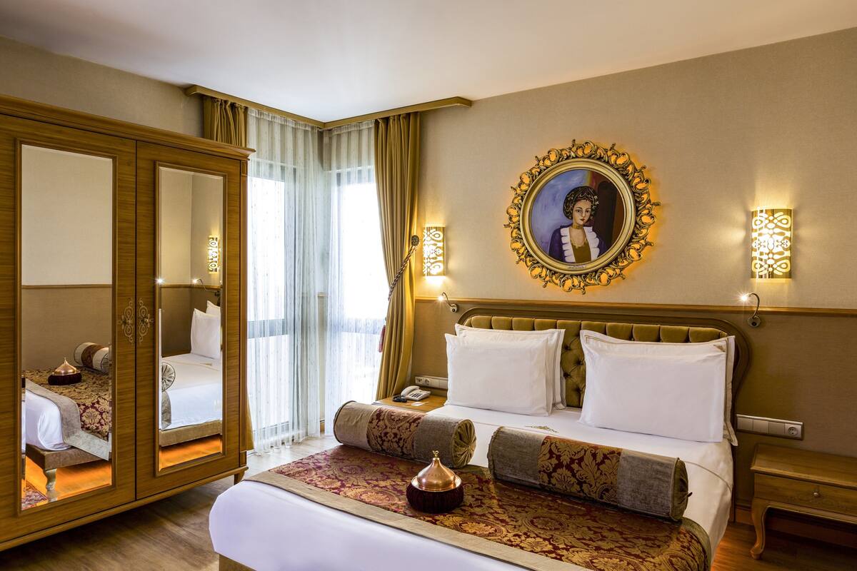 A hotel room with a large bed, a wardrobe with a mirror, a bedside table, a wall-mounted lamp, and a framed picture on the wall.