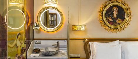 Une chambre d’hôtel avec un lavabo en marbre, un miroir rond, un lit avec du linge de maison blanc et un portrait encadré accroché au mur.