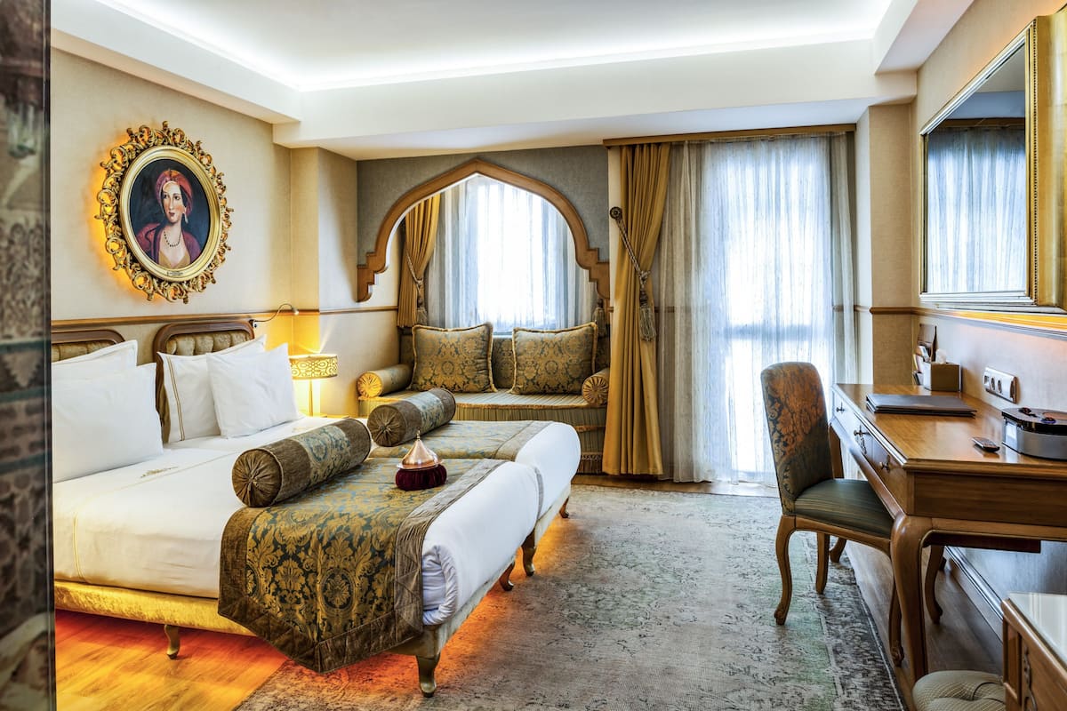 A hotel room with a large bed, a desk, and a chair. There is a painting on the wall and a window with curtains.