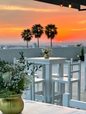 Una terraza en la azotea con mobiliario blanco, un florero con flores y vistas al atardecer.