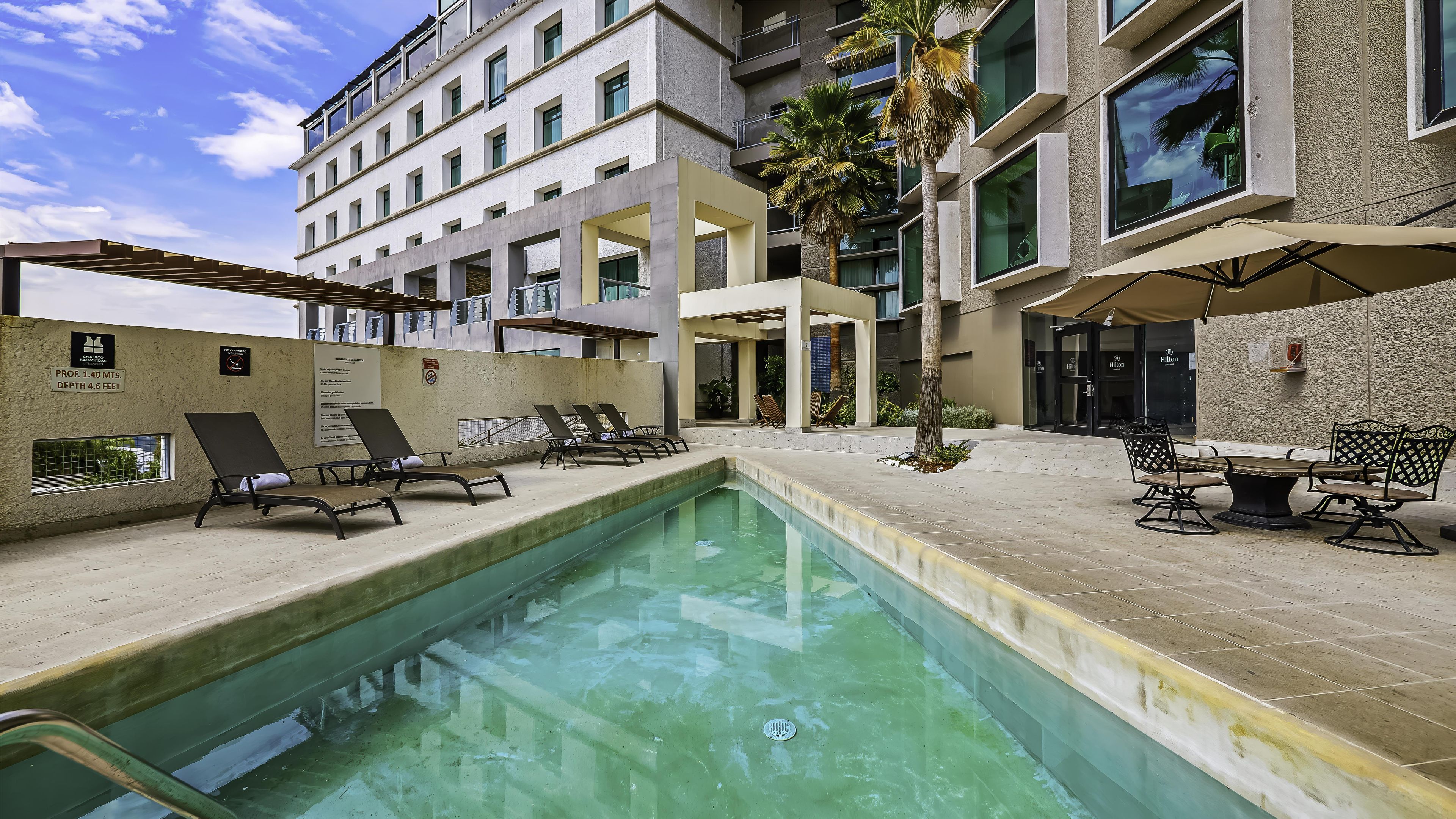 Una piscina con sillones de descanso y sombrillas, un edificio con varios pisos y balcones, y un letrero que indica una profundidad de 1,40 metros.