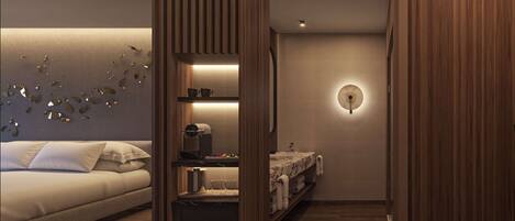 A modern hotel room with a bed, a built-in wardrobe, a small kitchenette, and a bathroom with a round mirror and wall-mounted light.