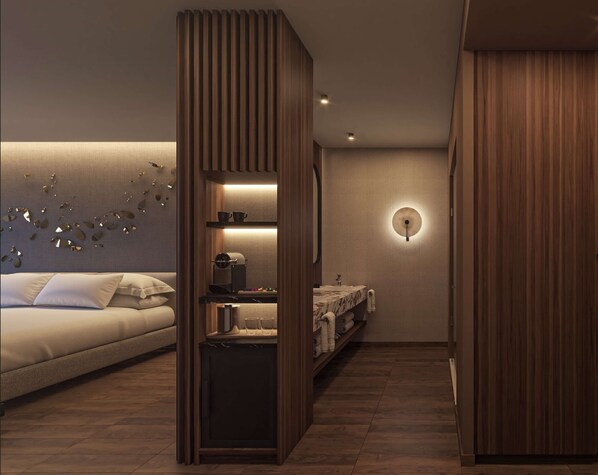 Una habitación de hotel moderna con una cama, armario empotrado, pequeña cocina y baño con un espejo redondo y luz en la pared.