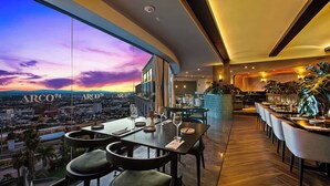 A restaurant with a city view, tables set with wine glasses, and a bar area with plants. - Hilton Queretaro (Querétaro)