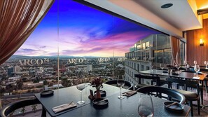 A restaurant with a city view, tables set with wine glasses and cutlery, and the name 'ARCO74' on the windows. - Hilton Queretaro (Querétaro)
