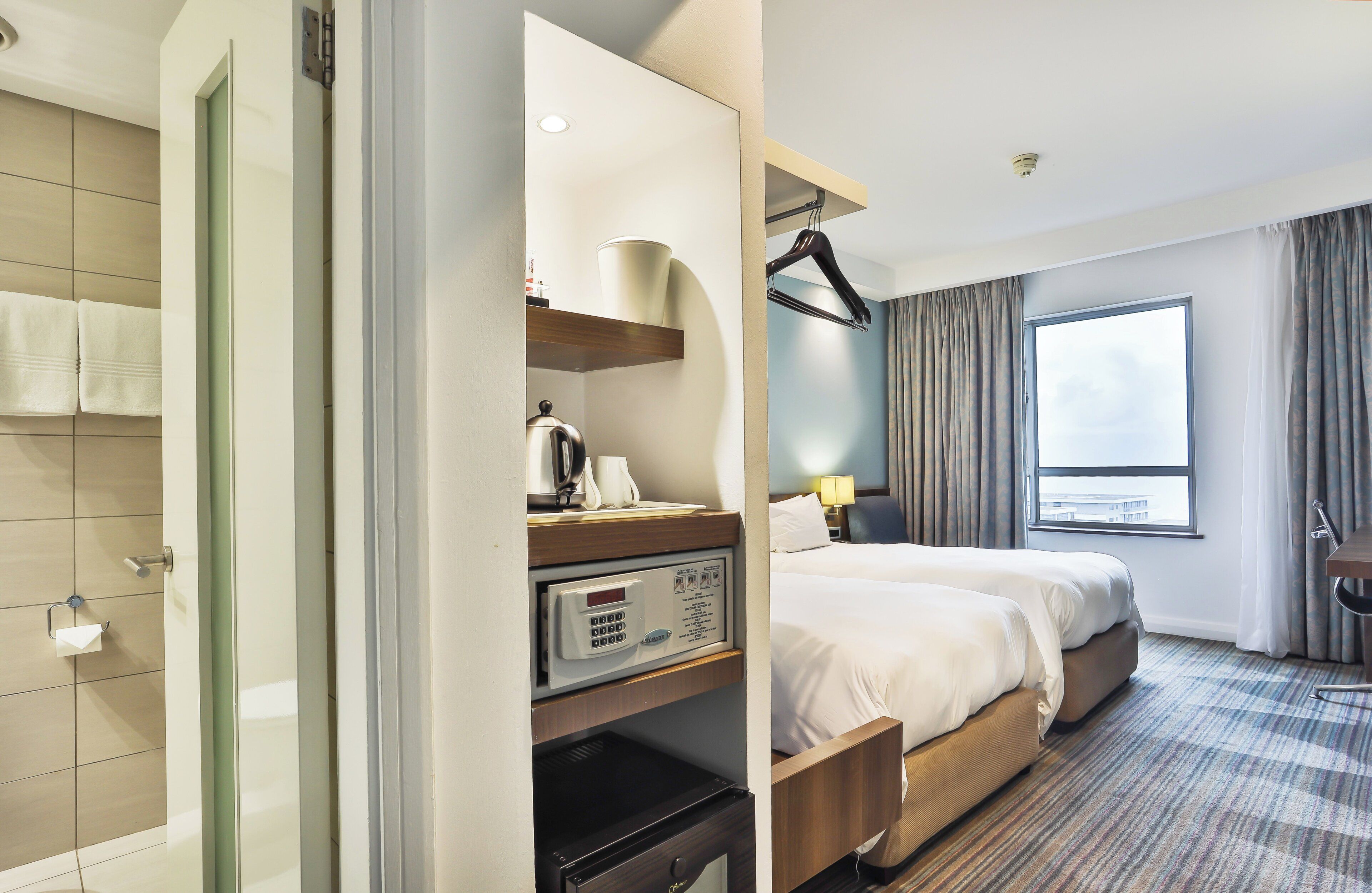 A hotel room with a bed, a bedside table with a lamp, a window with curtains, and a view of the city.