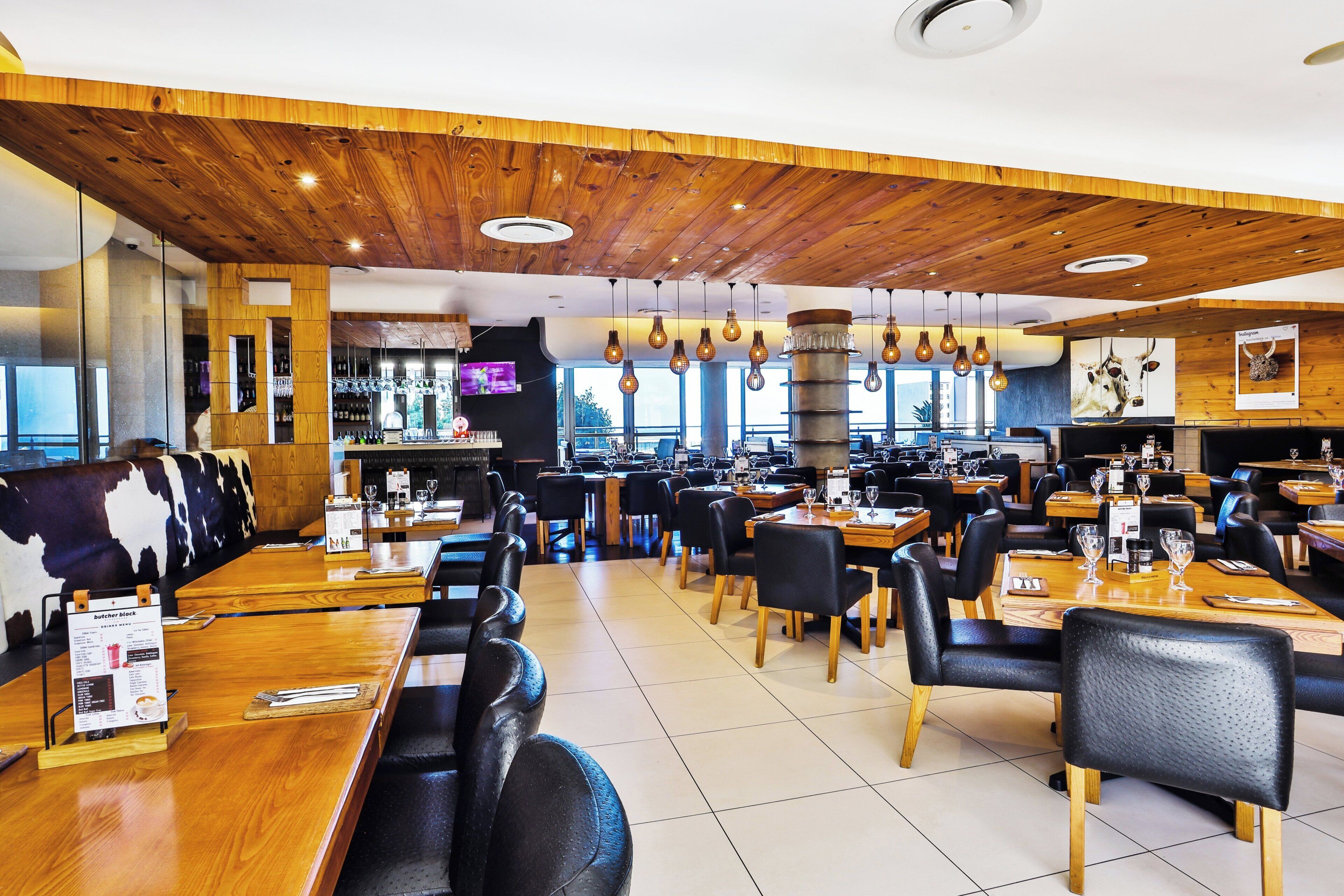 Restaurante con techo de madera, sillas de cuero negro y mesas dispuestas con copas de vino y menús.