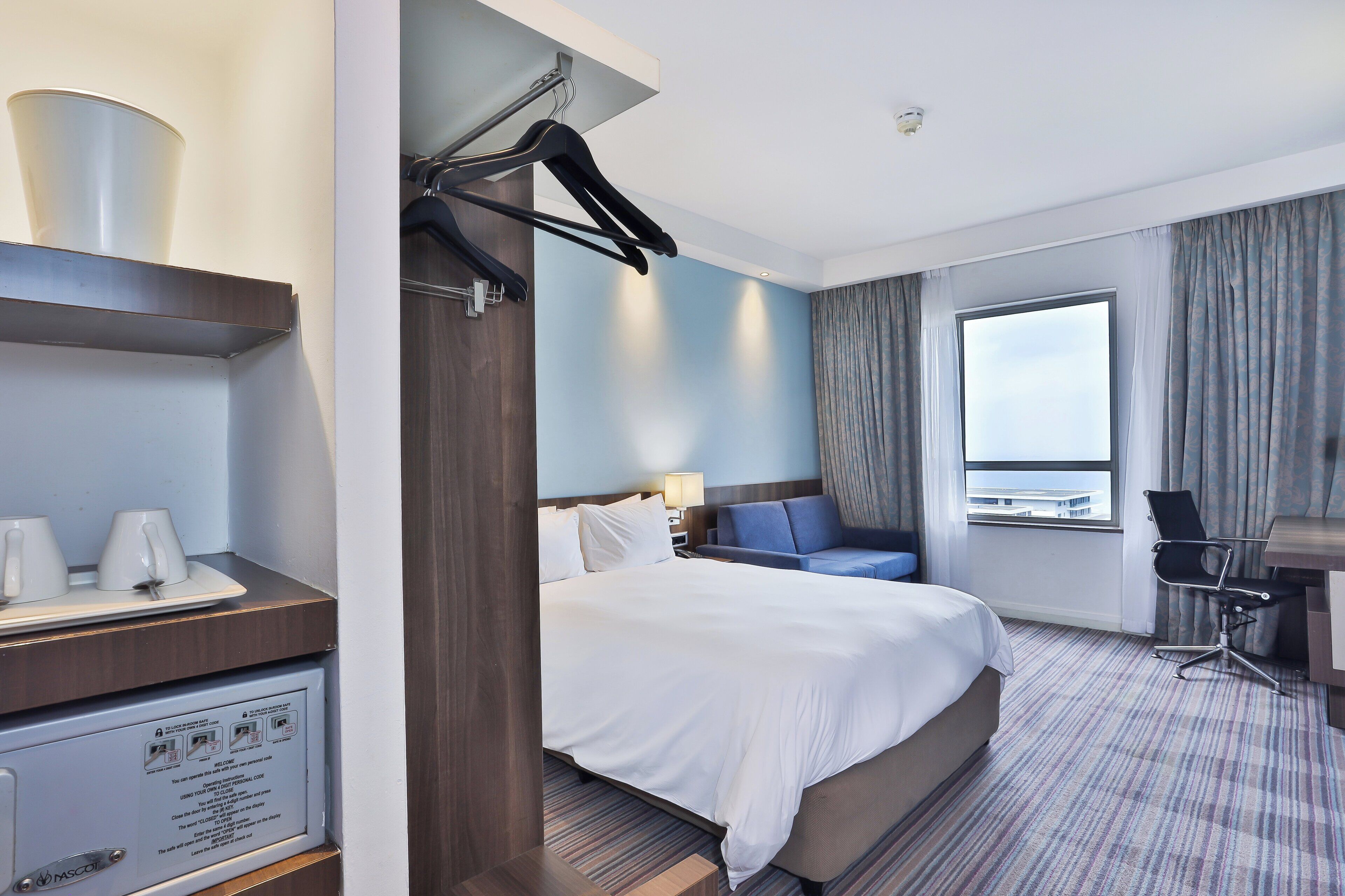 A hotel room with a bed, a desk, a chair, a window, and a minibar.