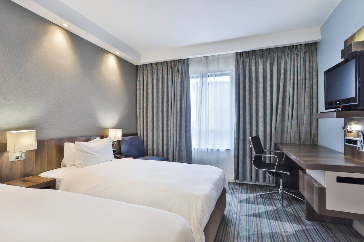 A hotel room with two beds, a desk, a chair, a television, and a window with curtains.