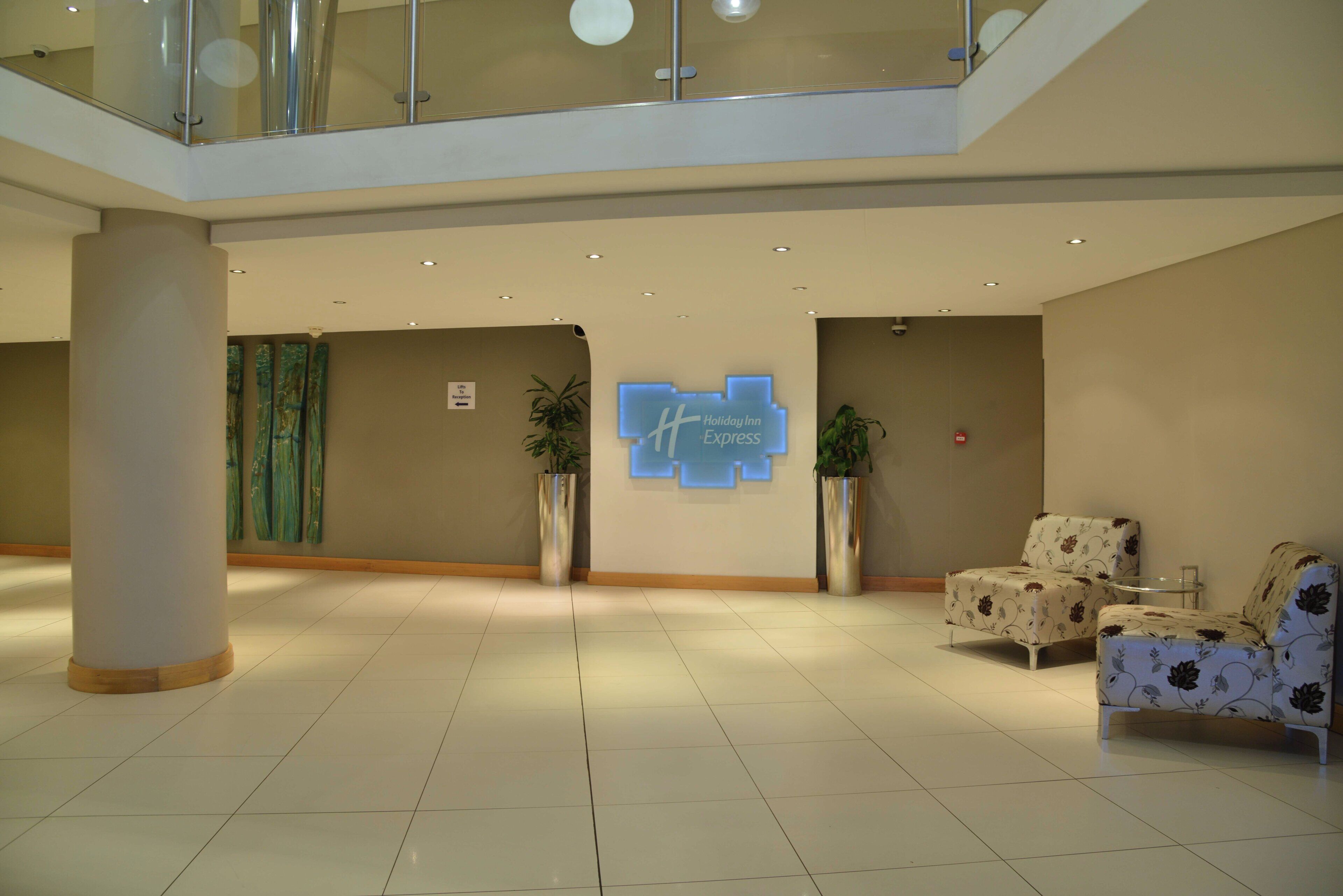 Un vestíbulo amplio con una mesa de recepción, dos plantas en macetas, una zona de estar con sillas de estampado floral y un letrero digital que indica Holiday Inn Express.