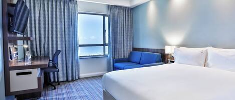 A hotel room with a bed, a desk with a chair, a TV, and a window with curtains.