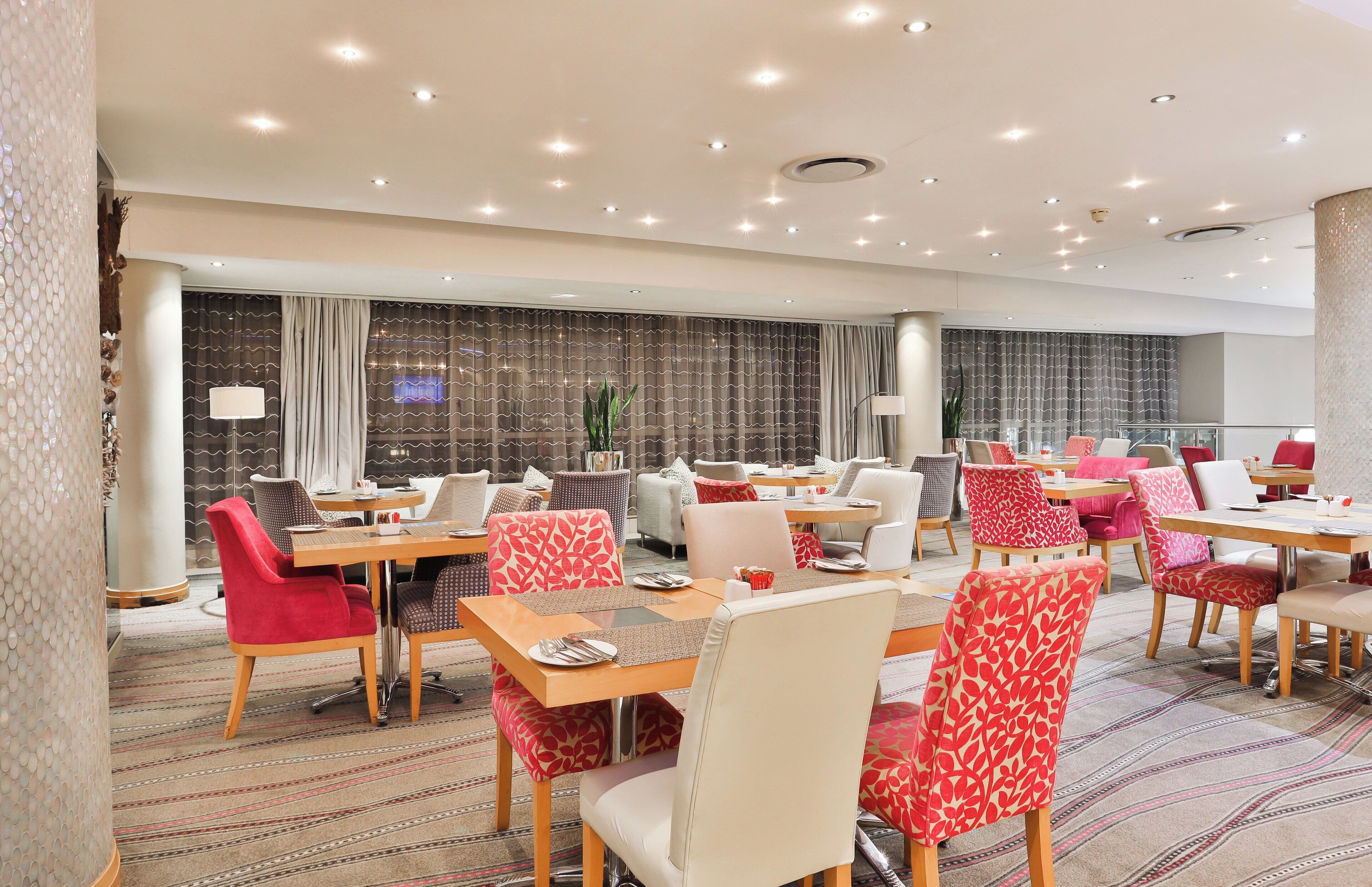 A hotel dining area with multiple tables and chairs, large windows with curtains, and a patterned carpet.