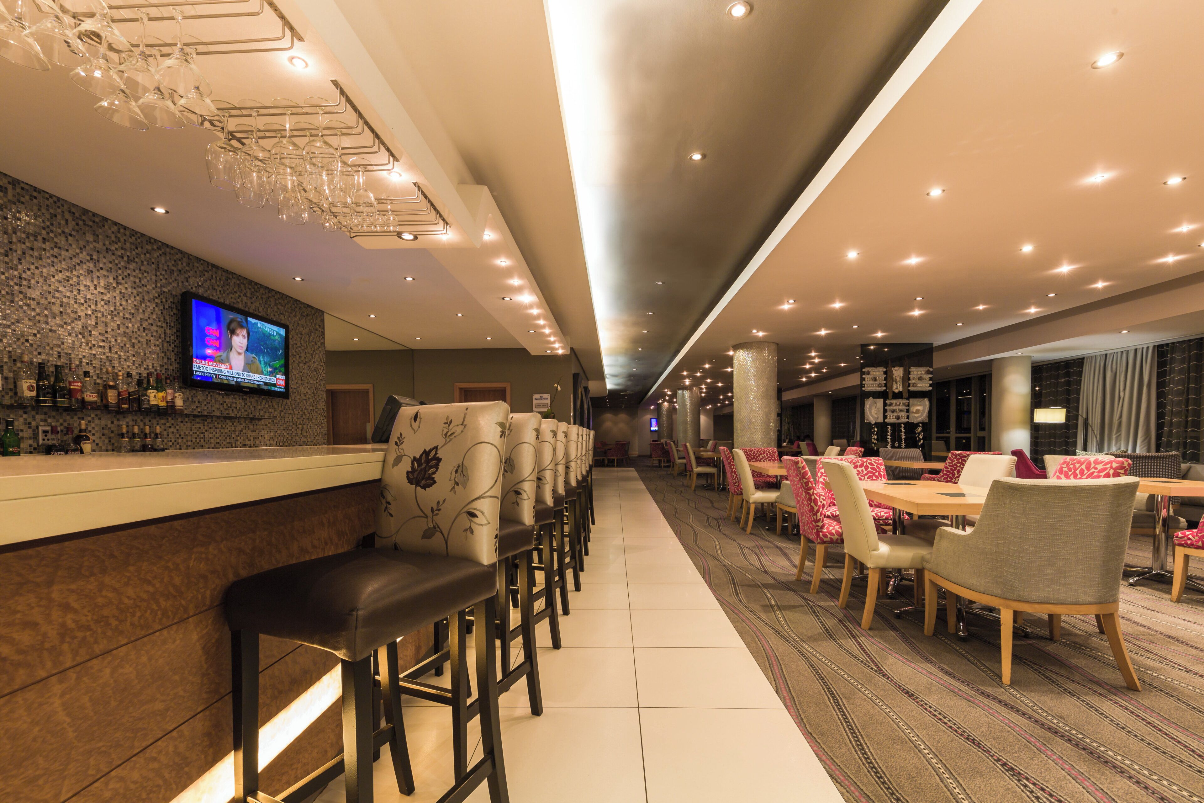 A hotel bar with a TV screen, bar stools, and a seating area with tables and chairs.