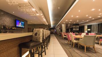 A hotel bar with a TV screen, bar stools, and a seating area with tables and chairs.