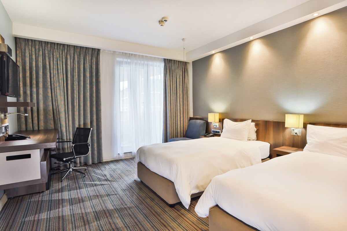 A hotel room with two beds, a desk with a chair, a television, and a window with curtains.