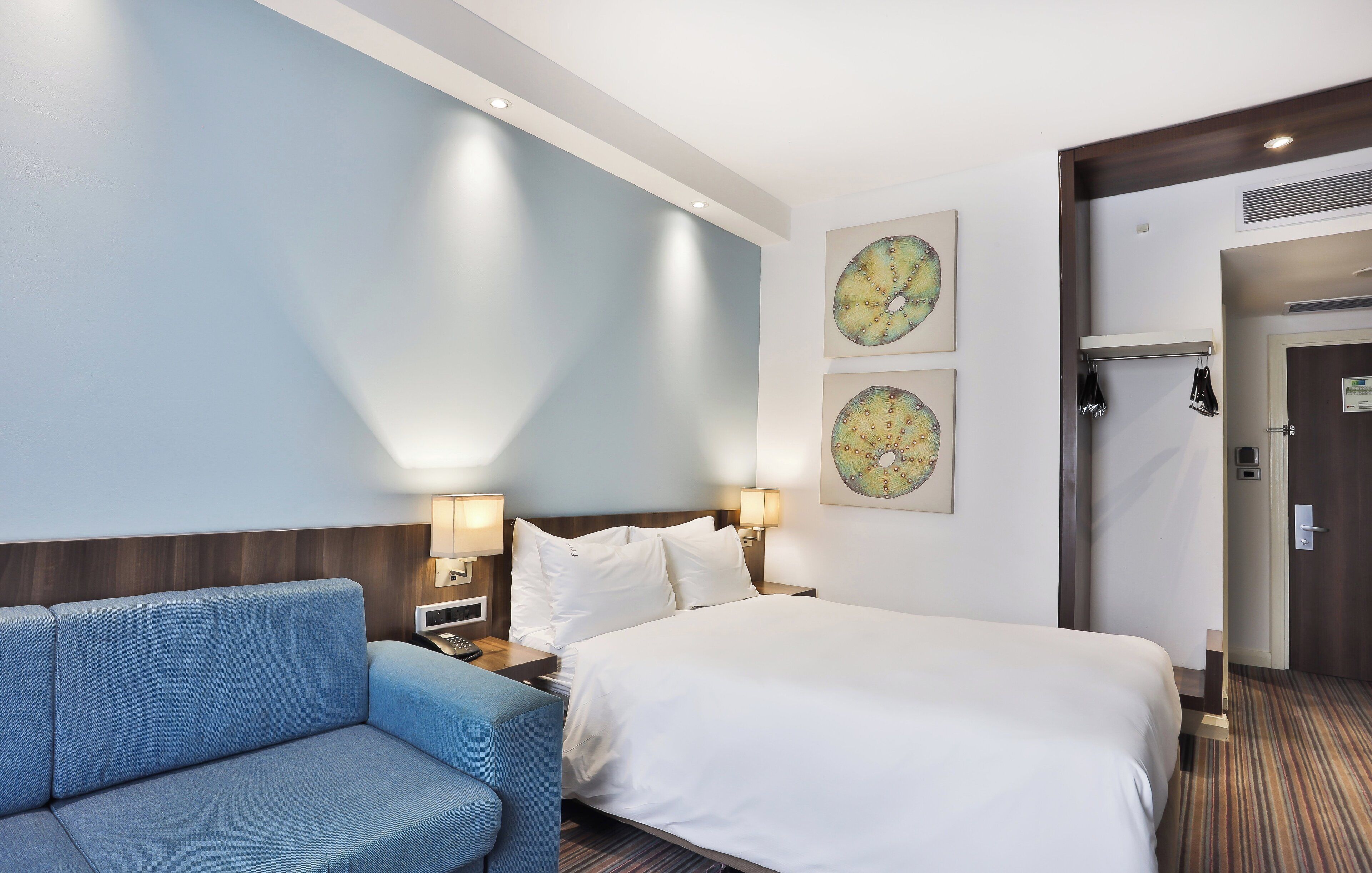 A hotel room with a bed, a blue sofa, two wall-mounted lamps, and two wall art pieces.