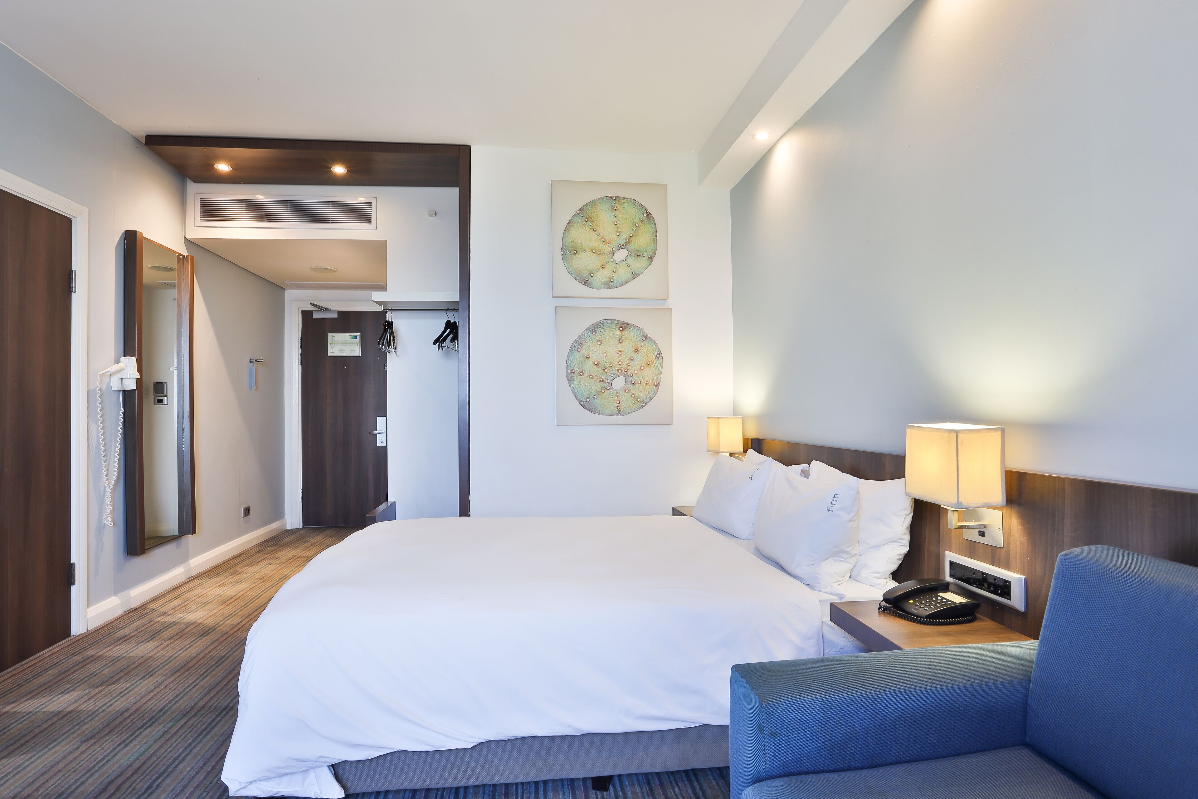 A hotel room with a large bed, a bedside table with a lamp, a chair, and two wall-mounted artwork pieces.