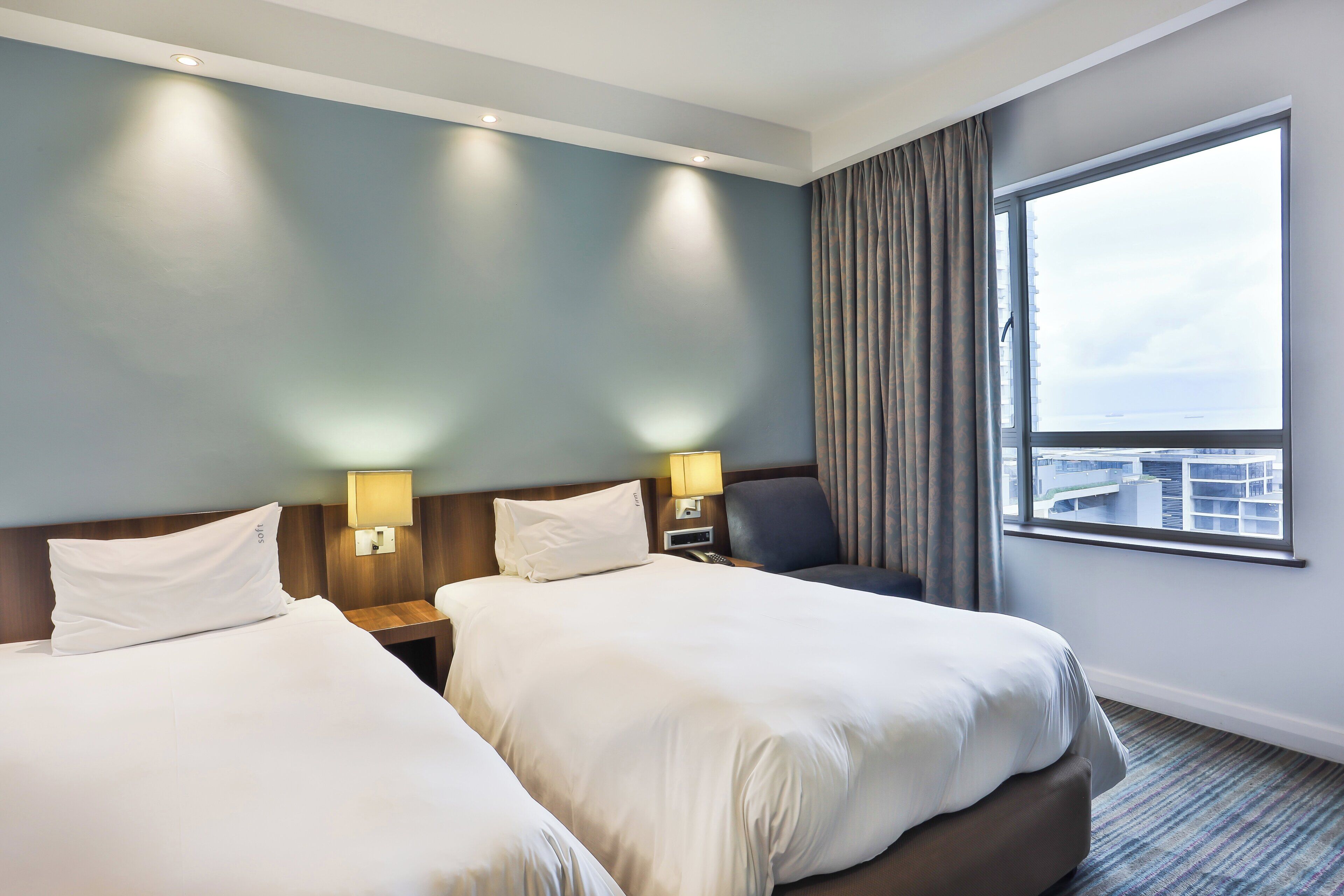 Une chambre d’hôtel avec deux lits, une table de chevet, une lampe, une chaise et une grande fenêtre donnant sur la ville.