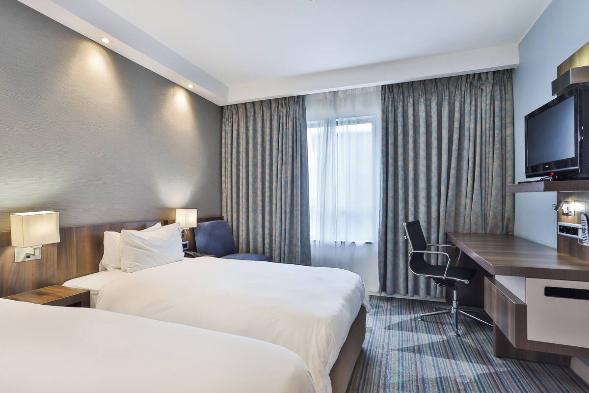 A hotel room with two beds, a desk, a chair, a television, and a window with curtains.