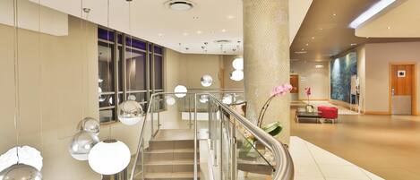 Un moderno vestíbulo de hotel con una escalera central, barandales de vidrio y lámparas decorativas.