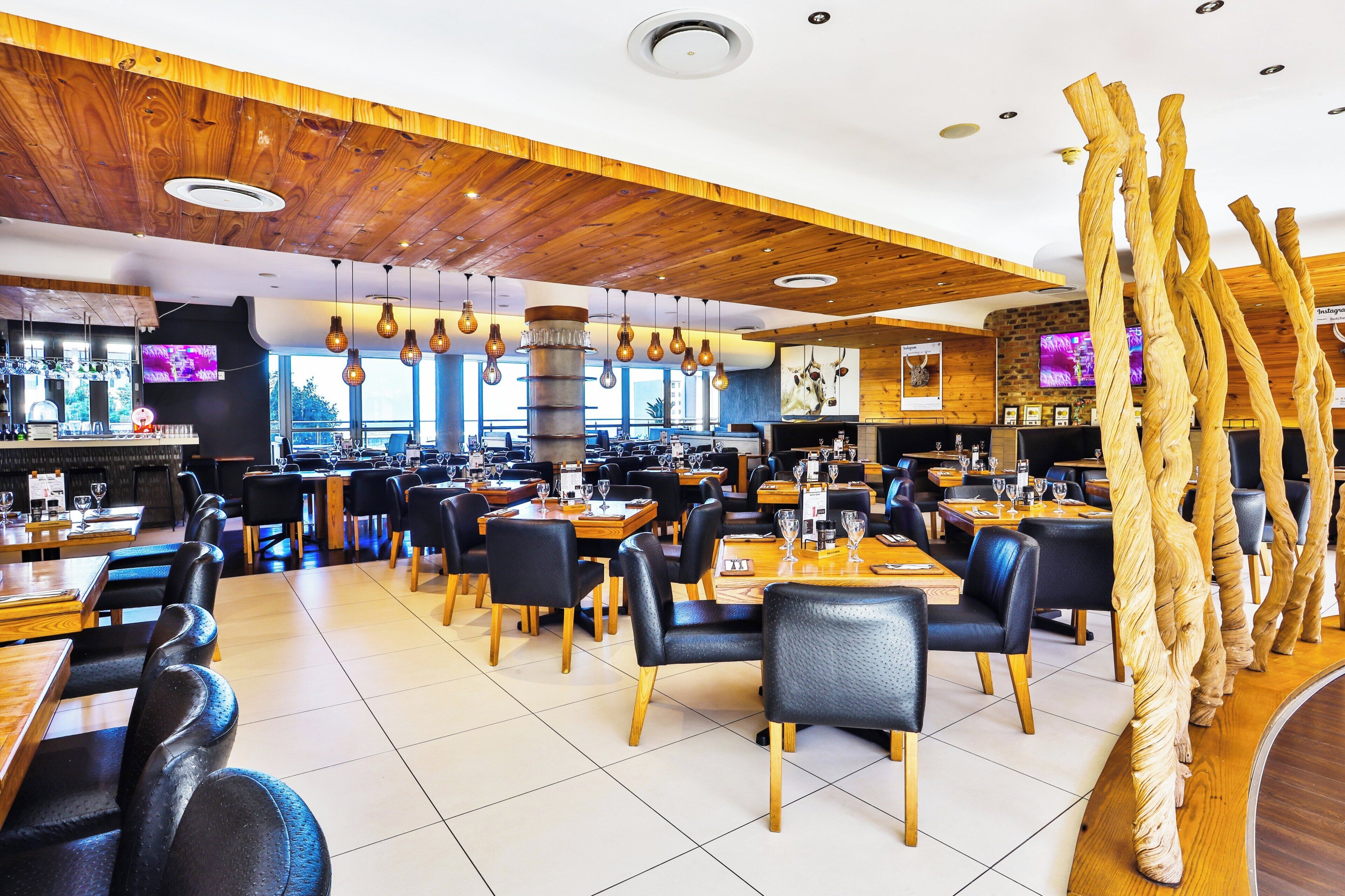 A restaurant interior with wooden ceiling, black leather chairs, and hanging light fixtures.
