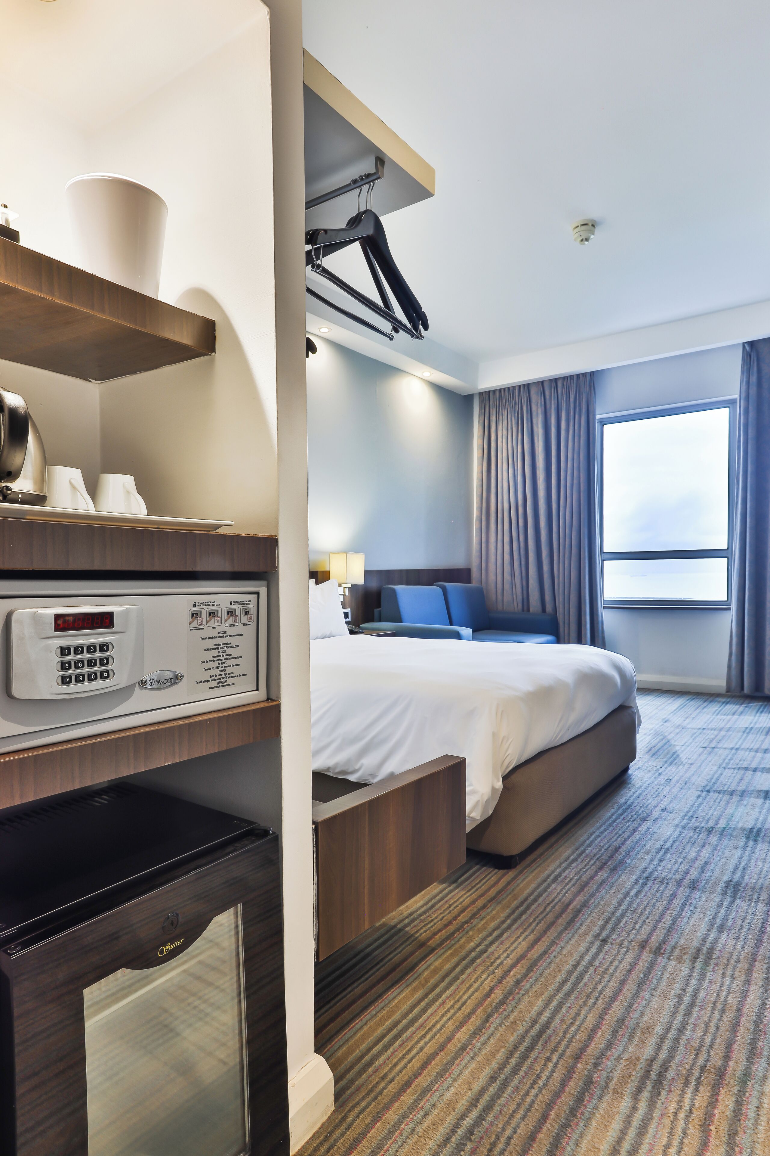 A hotel room with a bed, a desk with a computer, a coffee machine, and a window with curtains.