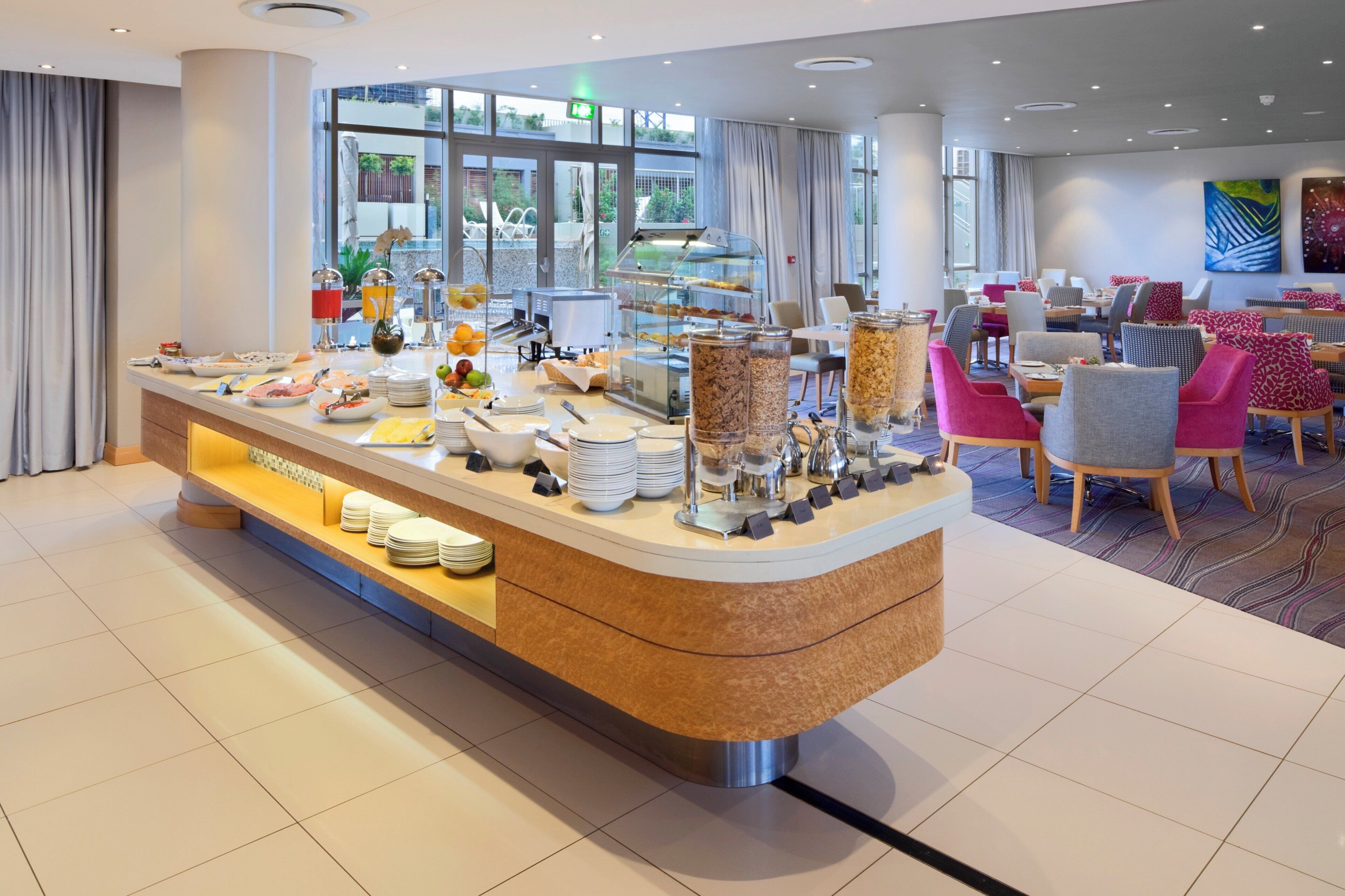 A modern breakfast buffet with a variety of food options, a seating area with colorful chairs, and a view of the outdoors through large windows.