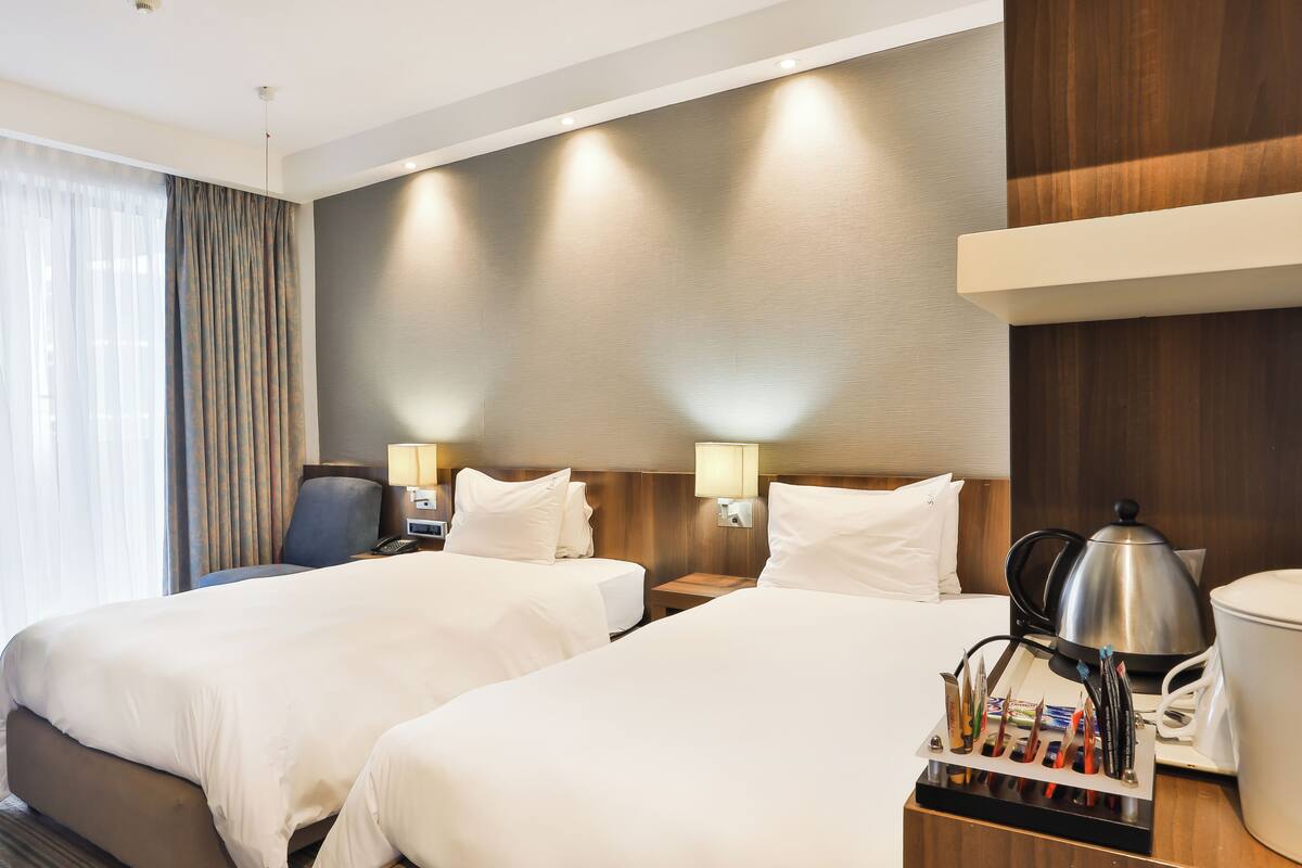A hotel room with two beds, a desk with a lamp, a coffee maker, and a view of the outside.