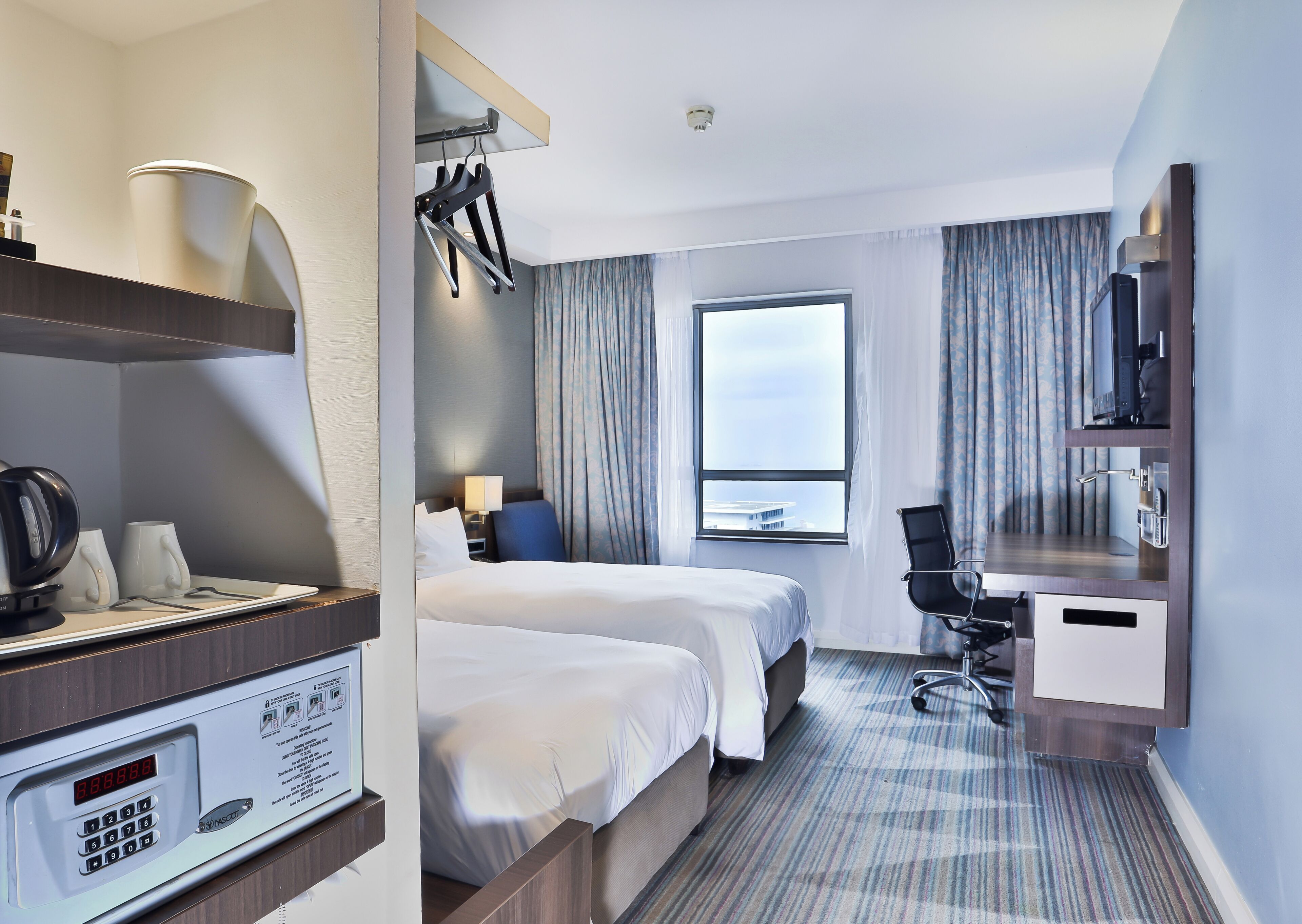 A hotel room with two beds, a desk, a chair, a TV, a window with a view, and a minibar.