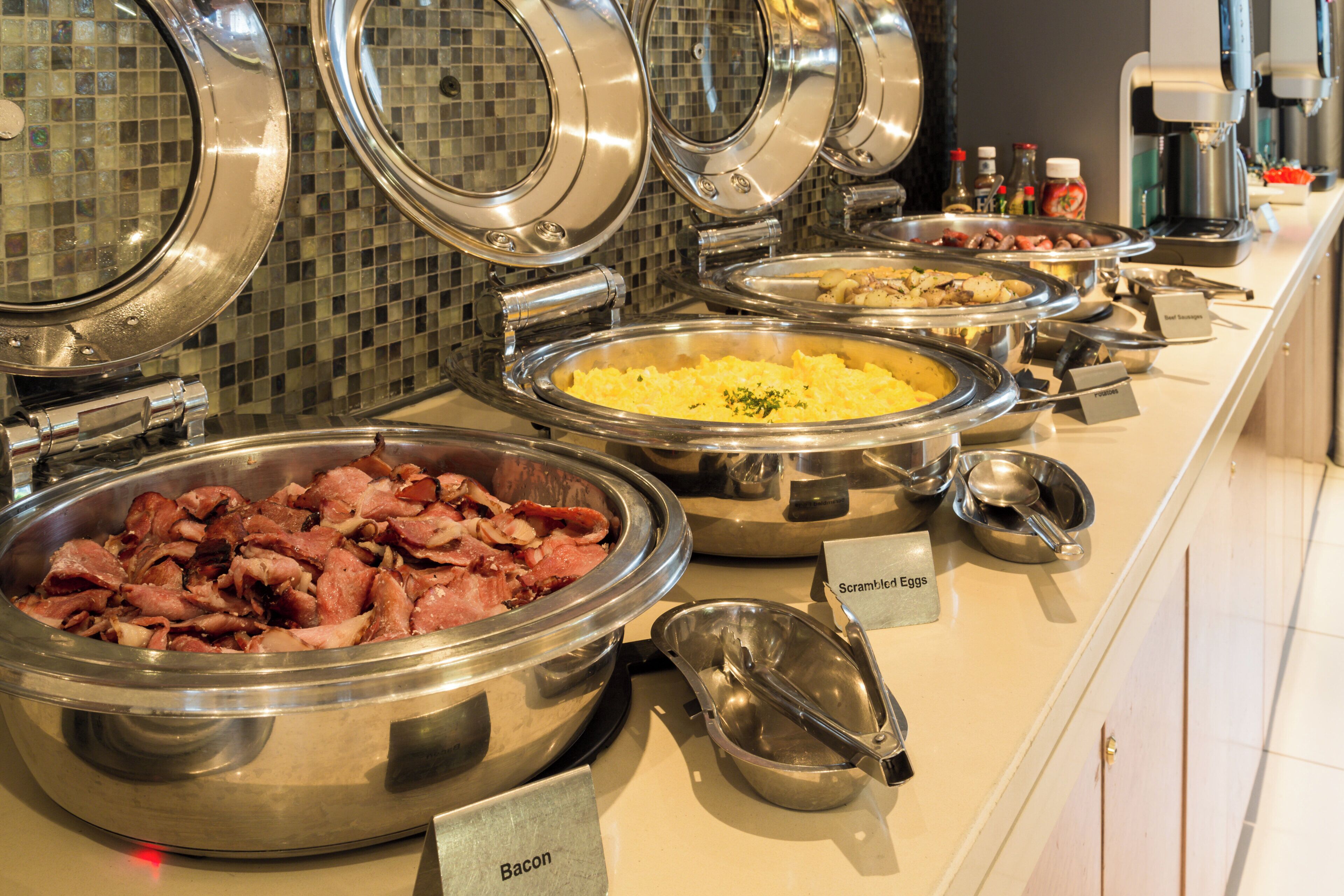Un bufé con diversas opciones de comida, incluyendo tocino, huevos revueltos y salchichas.