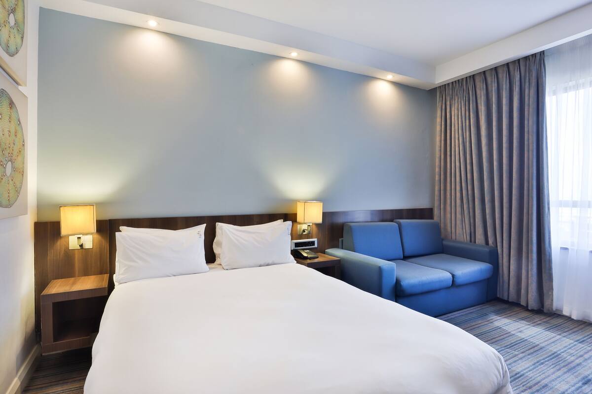 A hotel room with a large bed, two bedside tables with lamps, a blue armchair, and a view of the outdoors through a window with curtains.