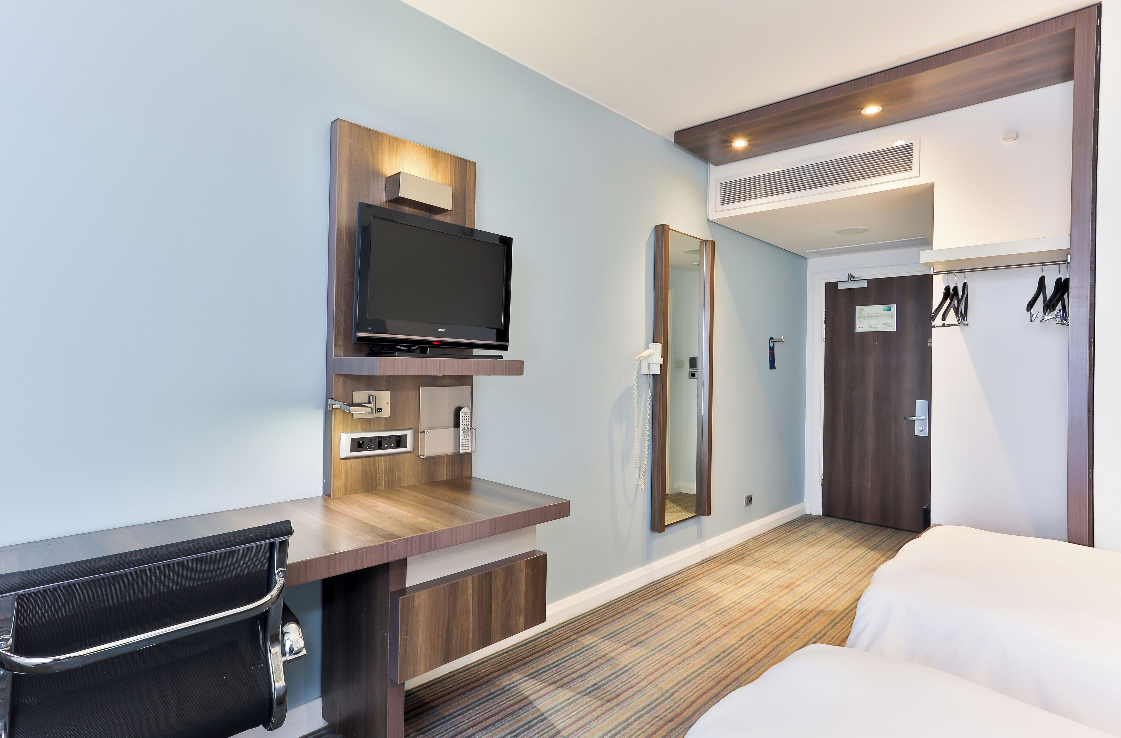 A hotel room with a flat-screen TV mounted on a wooden console, a mirror, a door with a coat rack, and a bed with white bedding.
