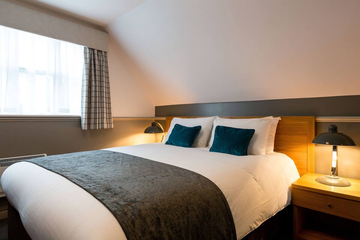 Un lit bien fait, avec une couverture grise, deux oreillers bleus et une tête de lit en bois. Une lampe de chevet se trouve à droite et une fenêtre avec des rideaux à gauche.