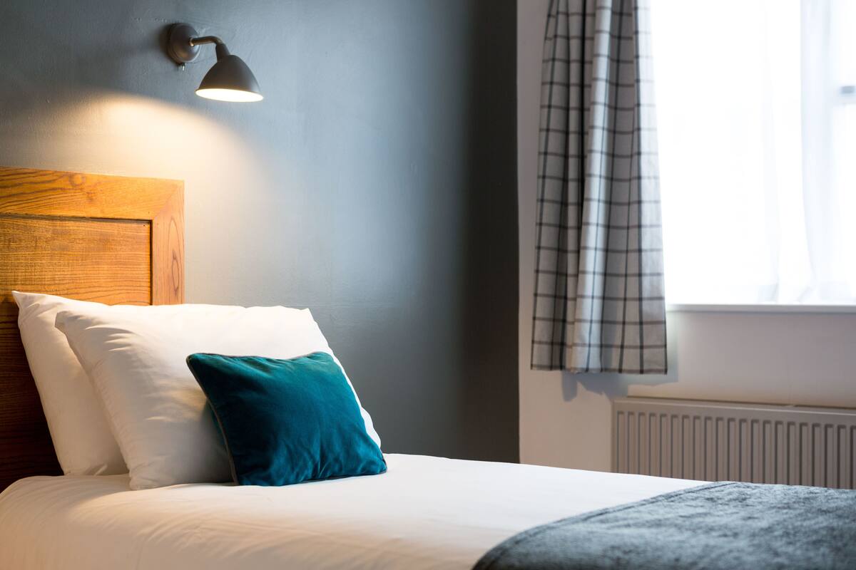 Un lit bien fait, avec des oreillers blancs et turquoise, une tête de lit en bois et un luminaire fixé au mur.