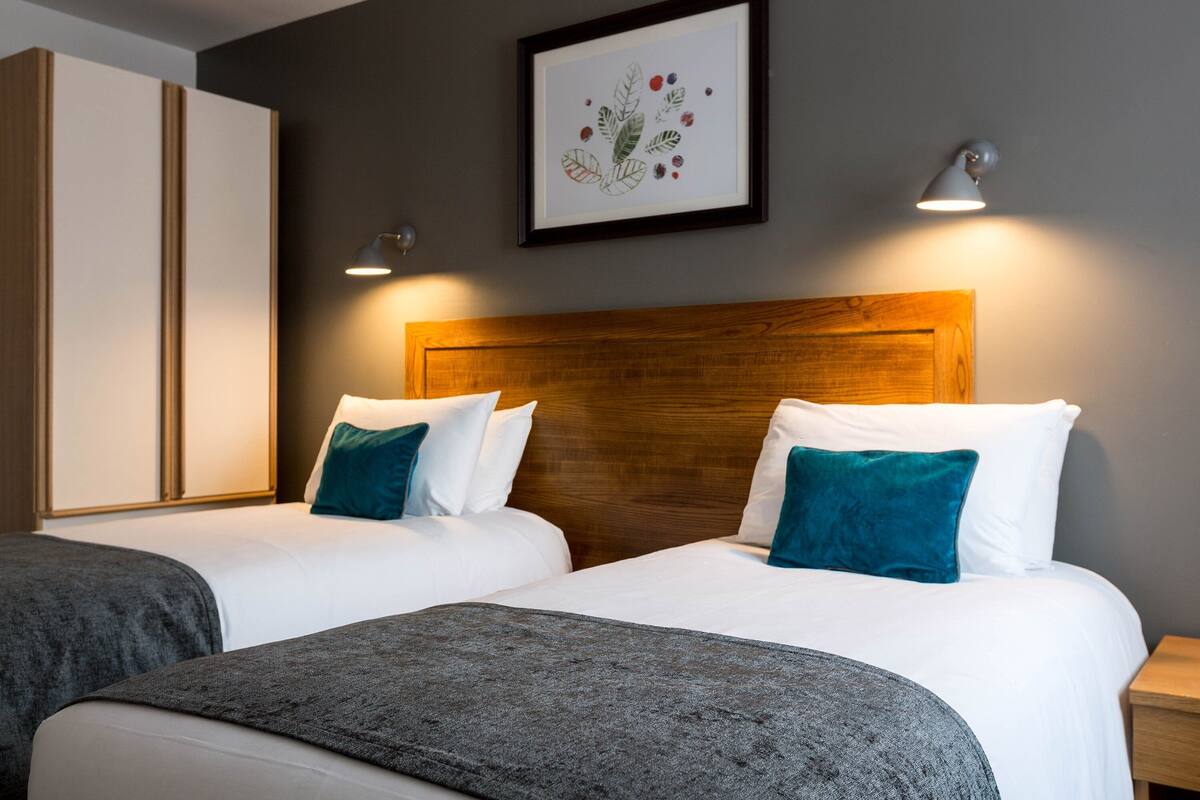 Une chambre d’hôtel avec deux lits, une tête de lit en bois, du linge de lit blanc, des coussins turquoise, un plaid gris, une estampe botanique encadrée au mur et deux lampes de chevet.